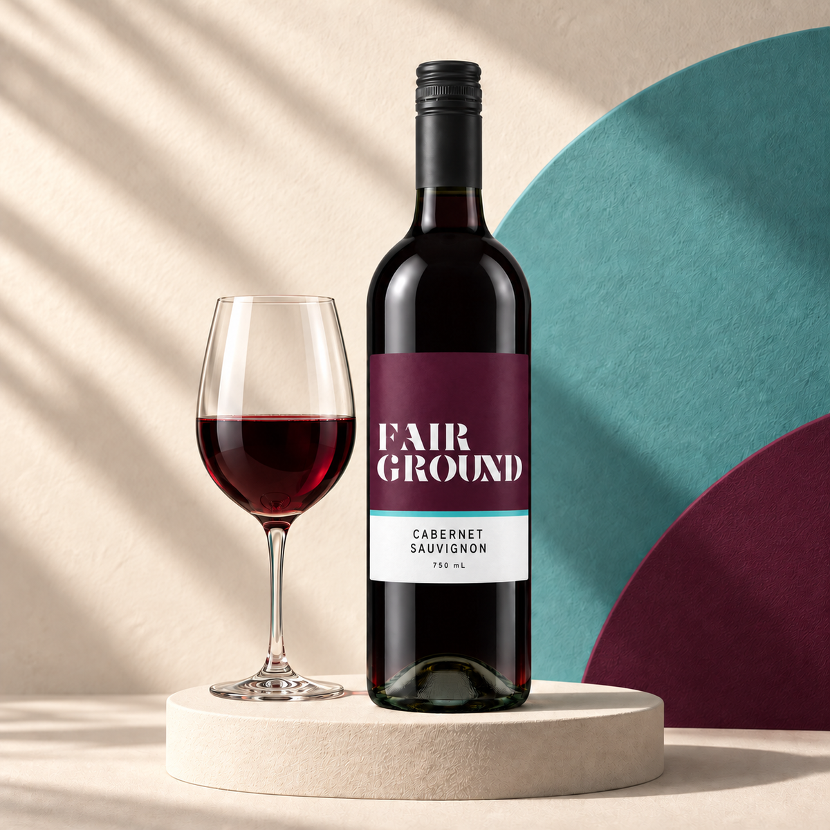 Fairground Cabernet Sauvignon bottle and a glass of red wine on a cream plinth with a teal and label-coloured backdrop