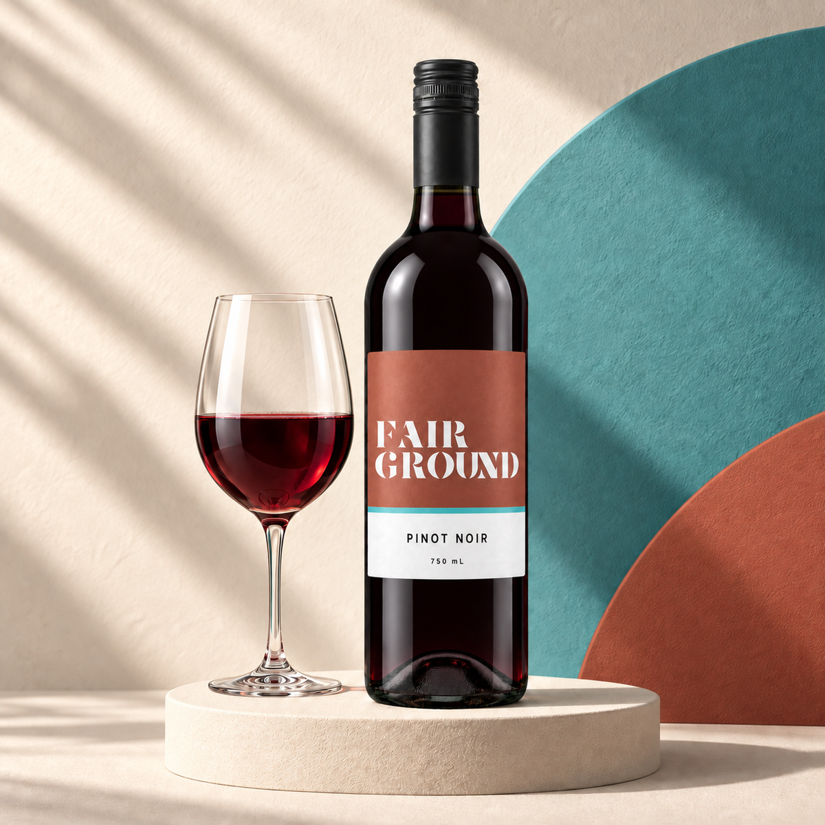 Fairground Pinot Noir bottle and a glass of red wine on a cream plinth with a teal and label-coloured backdrop